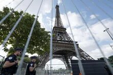 Eiffelturm mit Olympischen Ringen, bewacht von zwei Polizisten links. Bäume im Hintergrund. Absperrzaun im Vordergrund.