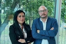 Foto: Eine Frau und ein Mann stehen nebeneinander vor einem Fenster. Die Frau links hat langes dunkles Haar und trägt ein schwarzes Jackett mit goldenen Knöpfen sowie eine weiße Bluse. Der Mann rechts hat eine Glatze, trägt eine Brille, ein blaues Sakko und ein weißes Hemd. Beide schauen freundlich in die Kamera und haben die Arme verschränkt.