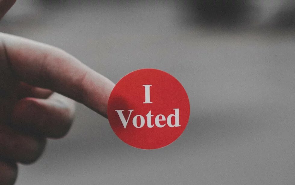 I Voted One hand is holding a sticker with "I Voted" written on it against a blurred background.