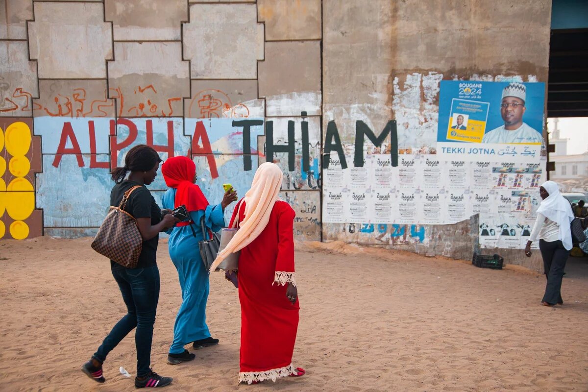 Wahlplakate in Dakar, Senegal, 2024. Drei Frauen laufen vor einer Wand mitr Wahplakaten lang.