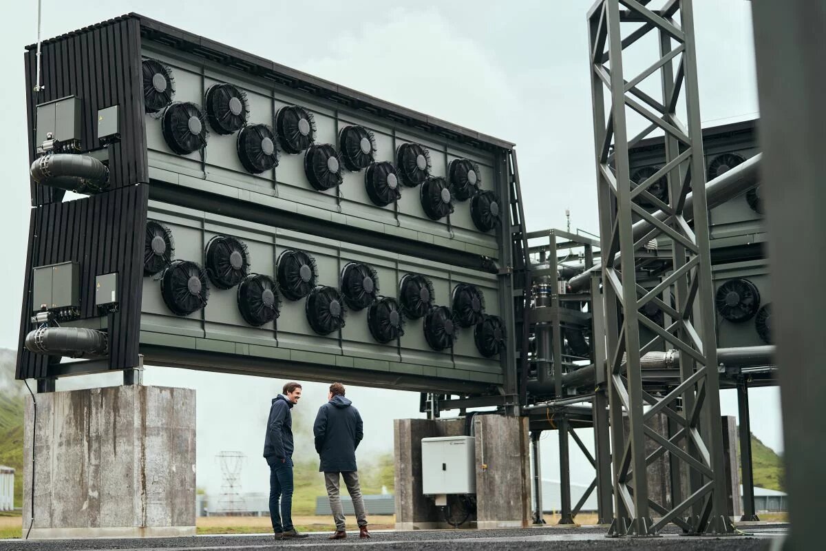 Direct Air Capture in Iceland Zwei Personen stehen vor einer großen, industriellen Anlage mit mehreren Lüftern. Stahlträger und Rohre sind sichtbar.