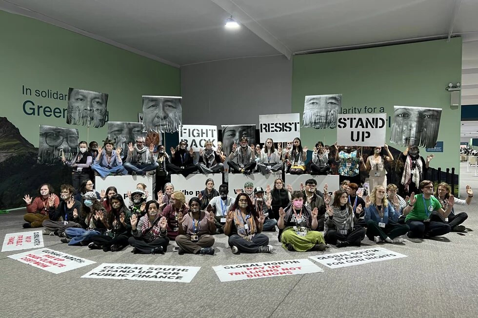 Klima-Aktivist*innen bei der COP29 in Baku Foto: Eine Gruppe Aktivist*innen sitzt und steht in einer Halle, hält Schilder mit Aufschriften wie „FIGHT BACK“ und „RESIST“ und hebt die Hände.
