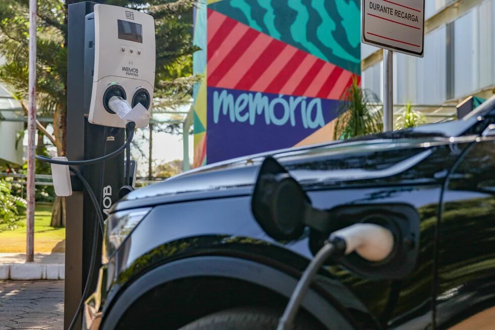 Elektroauto an einer Ladestation in Brasilia Foto: Ein Elektroauto lädt an einer Ladestation. Im Hintergrund eine bunte Wand mit "memória" und ein Schild mit Text.