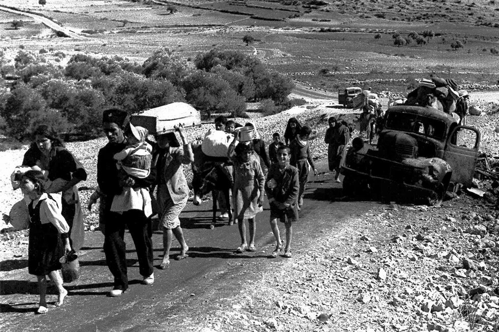 GPT Icon Photo: A group of people, including children, walk along a road carrying belongings. A destroyed truck and more people are in the background.